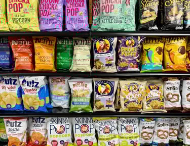 Round Hill Market snack aisle with various brands of popcorn, chips, and pretzels, including Boom Chicka Pop and Skinny Pop.