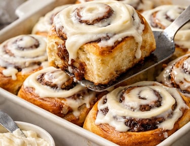 Freshly baked homemade cinnamon rolls with cream cheese frosting being served from a ceramic baking dish.