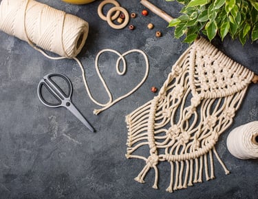 Tapiz de macramé hecho a mano con cordón de algodón, aros de madera y tijeras sobre una mesa oscura.