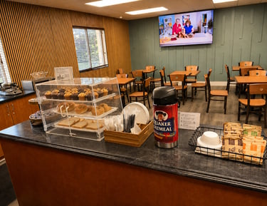 Hotel breakfast bar featuring fresh muffins, bagels, and a Quaker Oatmeal station in a dining area.