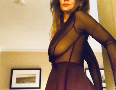 A woman posing in a dark sheer long-sleeve top and matching skirt in a room.