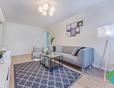 Salon lumineux avec un canapé gris clair, un grand tapis gris foncé, une table basse en verre et un meuble télé blanc