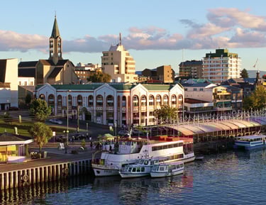 Valdivia city waterfront with tour boats docked at the pier near historic buildings and a church spire.