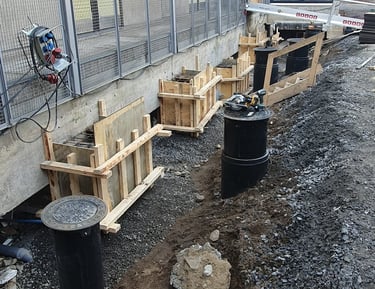 Foundation construction with wooden formwork and black drainage pipes next to a modern building.