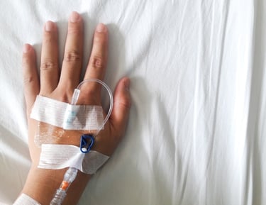 Patient hand with a medical IV drip catheter and bandages on a white hospital bed sheet.