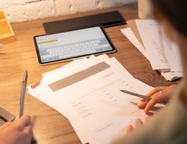 imagem de folhas com descrções de itens, tablet ao fundo e mãos de um homem trabalhando e fazendo anaotações, sobre mesa