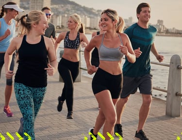 A diverse group of smiling friends jogging along a scenic coastal boardwalk at sunset.