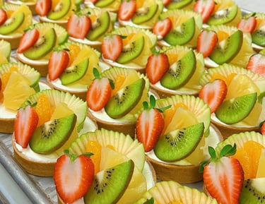 Fresh fruit tarts topped with sliced strawberries, kiwi, and orange on a bakery tray.