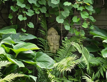 A stone Buddha head statue nestled among lush green garden ferns, hostas, and grape ivy leaves.