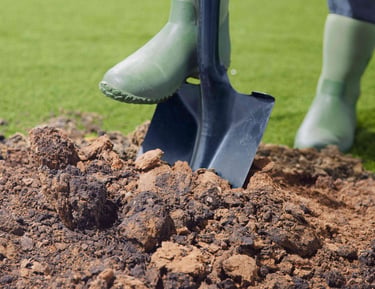A gardener in green rubber boots uses a metal shovel to dig into a pile of garden soil on a lawn.