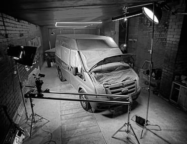 Filmavimo aikštelė garaže, kurioje yra furgonas su studijiniu apšvietimu ir kameros slideriu.