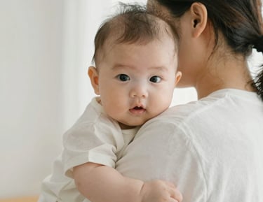 A baby is peacefully sleeping on a light blue textured blanket, wearing pajamas with a cute animal print. The child is wrapped in a soft, gray hooded blanket. A pacifier is gently held in the sleeping baby's mouth. Red rose petals are scattered beside the baby on the bedding.