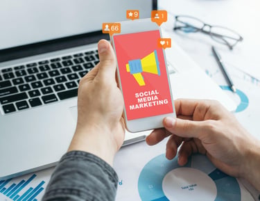 A person holding a smartphone displaying a social media marketing graphic next to a laptop and data charts.