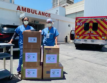 Two healthcare workers in scrubs and masks standing beside stacked supply boxes labeled with heart l