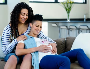 A happy lesbian couple sitting on a sofa together while touching a pregnant belly with baby shoes.