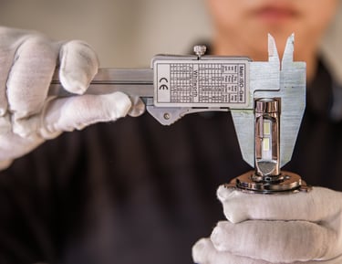 A technician in white gloves uses a metal vernier caliper to measure an automotive LED headlight bulb.