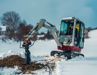 Excavator digging a post hole through frozen ground to reach the frost line for structural stability