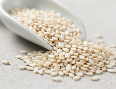 Extreme macro close-up of white sesame seeds spilling from a ceramic scoop onto a pale stone surface, bright diffused daylight, individual seed texture and hull detail fully visible, shallow depth of field, no human subjects