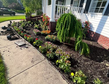 A neatly landscaped garden bed with colorful flowers and mulch along a white house porch and sidewalk.