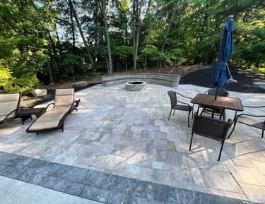 Custom stone paver patio featuring a round fire pit, built-in curved bench, and outdoor furniture in a wooded backyard.