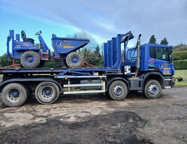 A blue flatbed transport truck carrying a Thwaites site dumper for construction equipment delivery.
