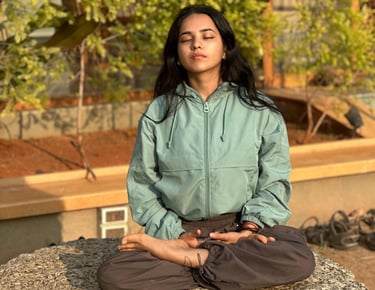 A young girl practicing mindful meditation in a lotus yoga pose on a large rock outdoors.