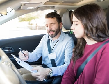 A professional driving instructor with a clipboard giving a student driver feedback inside a car.