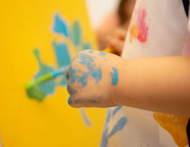Child's hand covered in blue paint while creating artwork on a yellow canvas