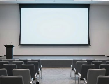 Clean photography of a modern event hall in Minneapolis. A minimalist stage is set with a podium and a large blank projection screen. The seating is arranged for a tech talk, with sleek contemporary chairs. Lighting is crisp and cool, utilizing a light gray and slate blue color scheme. North American / US.