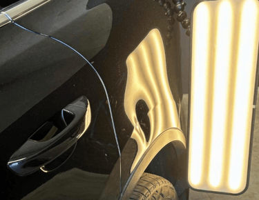 Professional paintless dent repair on a black car using a specialized LED light board to identify door dings.