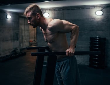 Muscular man performing triceps dips on parallel bars in a dark industrial gym setting.