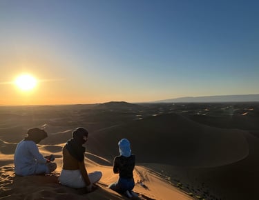View from behind as three people sit atop a sand dune at sunset on a Moroccan Sahara trek