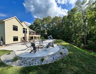Modern backyard concrete patio with a fire pit, Adirondack chairs, and a swimming pool slide near lush green trees.