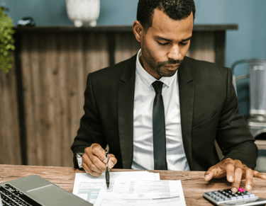 Homme en costume assis à son bureau, tenant un stylo et utilisant une calculatrice