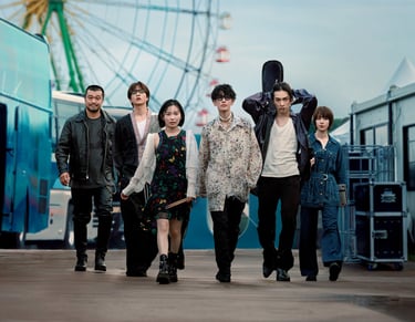 A stylish Japanese rock band walking outdoors past a tour bus and Ferris wheel at a festival venue.