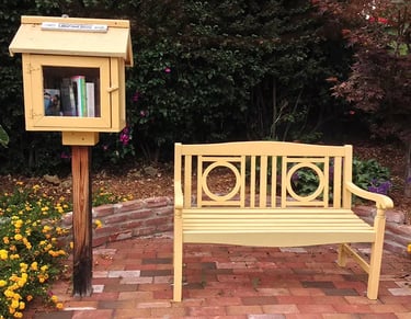 Yellow Little Free Library box on a post next to a matching yellow wooden garden bench on a brick patio.