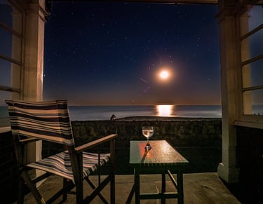 view of the moon over the sea as seen from a beach hut. A deck chair and glass on a table too