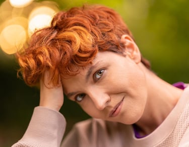 Portrait of a person with curly red hair and hazel eyes posing outdoors with soft bokeh lighting.