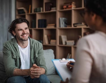 A smiling man sits on a sofa during a mental health therapy session with a counselor.