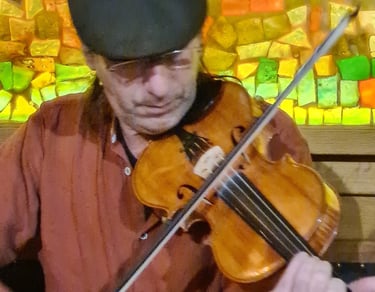 A musician playing a wooden fiddle in front of a colorful mosaic background at a live music pub.