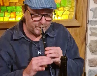 A man wearing a flat cap and glasses plays a traditional Irish tin whistle in a pub.
