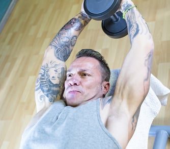 A man with arm tattoos performs a dumbbell overhead triceps extension on a weight bench at the gym.