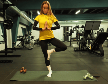 Una donna in posa di yoga albero (Vrksasana) su un tappetino da yoga in palestra. Indossa una maglietta gialla con logo 'Move