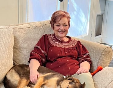 Photo of Debbie, seated on the couch with a resting medium-sized, brown and tan dog.
