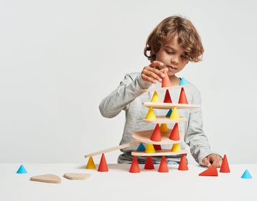 A boy creating a structure from red, blue and yellow silicone cones and wooden tables in the room