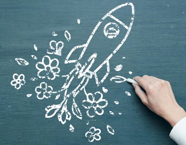 A hand holding white chalk draws a rocket propelled by flowers on a chalkboard.