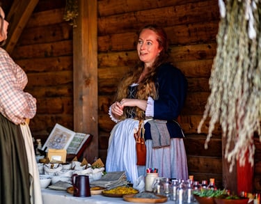 a woman in 18th century clothing stands behind a table covered in herbal remedies