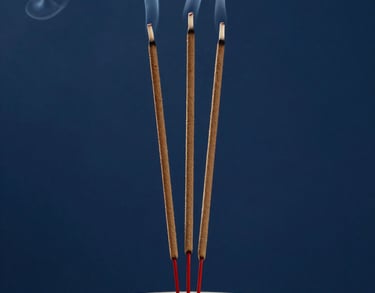 A close-up of incense sticks burning in a ceramic holder, with soft smoke curling upwards against an indigo background, South Asian / Tamil Nadu, India.