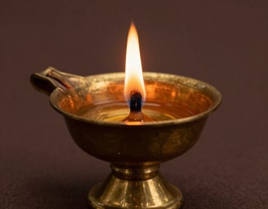 A close-up photograph of a sacred brass oil lamp with a single flickering flame, casting a warm golden glow against a dark brown background. South Asian spiritual aesthetic.