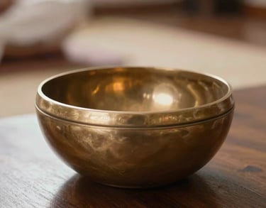 A close-up photograph of a brass singing bowl reflecting a soft amber glow on a dark wooden table in a serene meditation room, South Asian / Tamil Nadu, India.